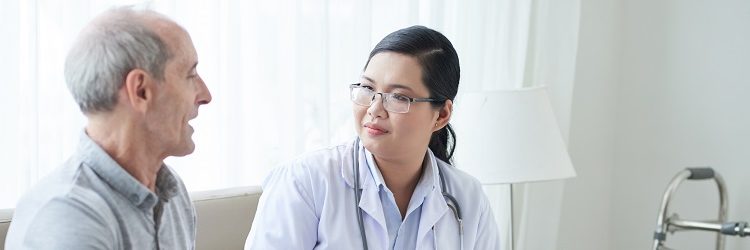Asian adult woman with clipboard sitting on sofa in house of senior man during visit and listening to patient