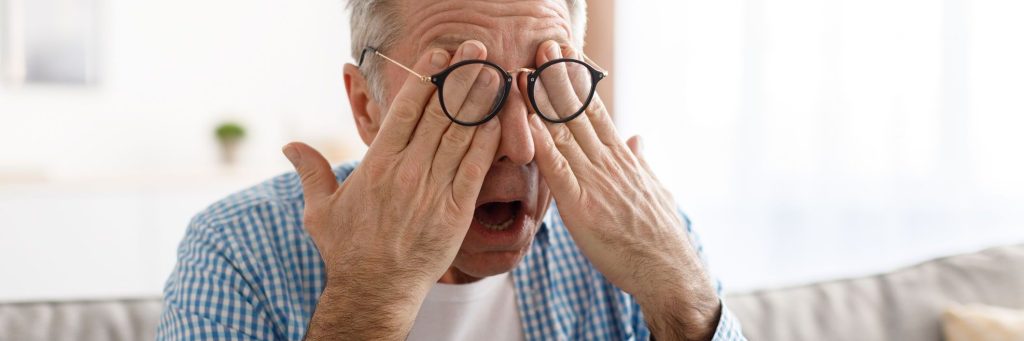 Glaucoma. Senior Man Rubbing Tired Eyes Wearing Eyeglasses Having Poor Eyesight Feeling Suffering From Eyestrain Sitting On Couch At Home. Ophtalmology